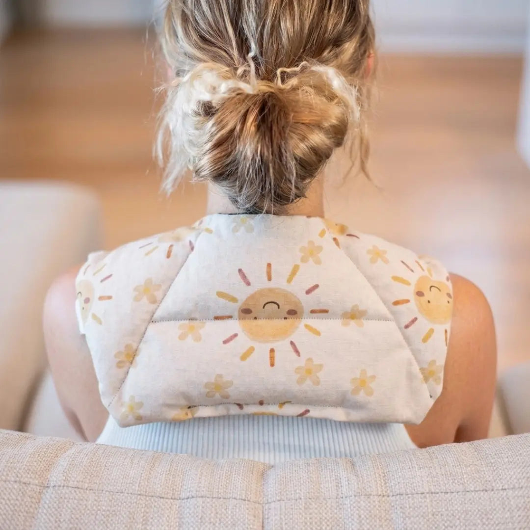 A woman viewed from behind sitting on a couch, wearing Happy sun fabric neck heat pack filled with lupin seeds, worn across the shoulders for hot or cold therapy