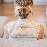 A woman viewed from behind sitting on a couch, wearing Happy sun fabric neck heat pack filled with lupin seeds, worn across the shoulders for hot or cold therapy